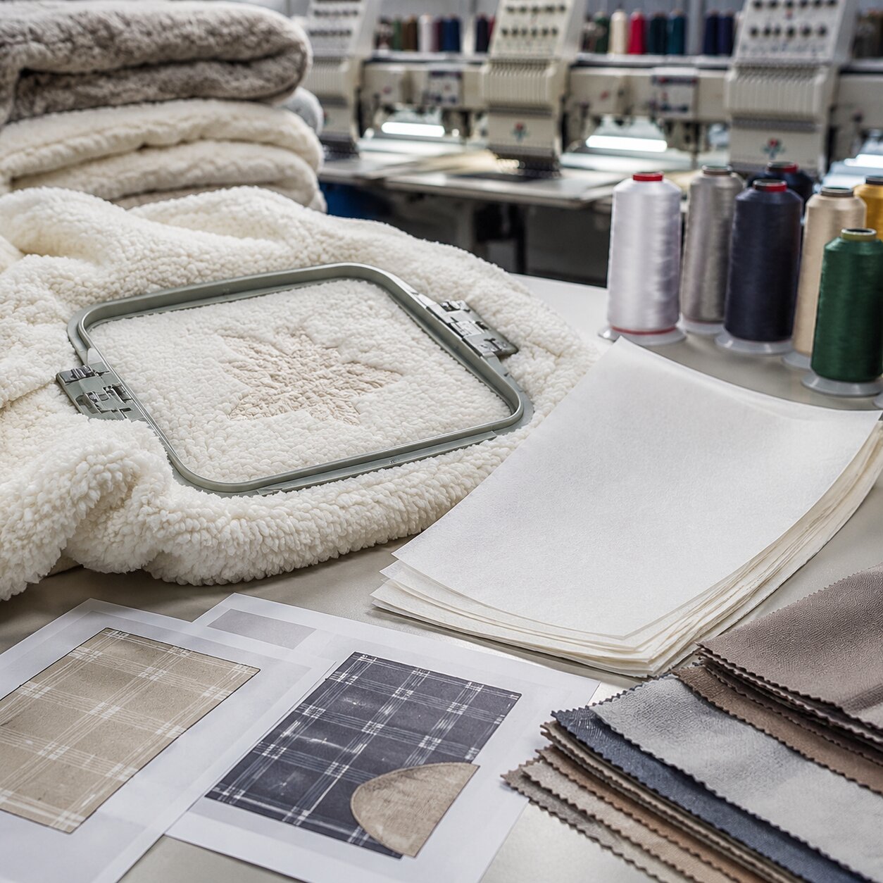 Close-up of a 300gsm sherpa blanket being hooped and embroidered in a factory QC area with stitch samples, thread cones, and approval swatches nearby